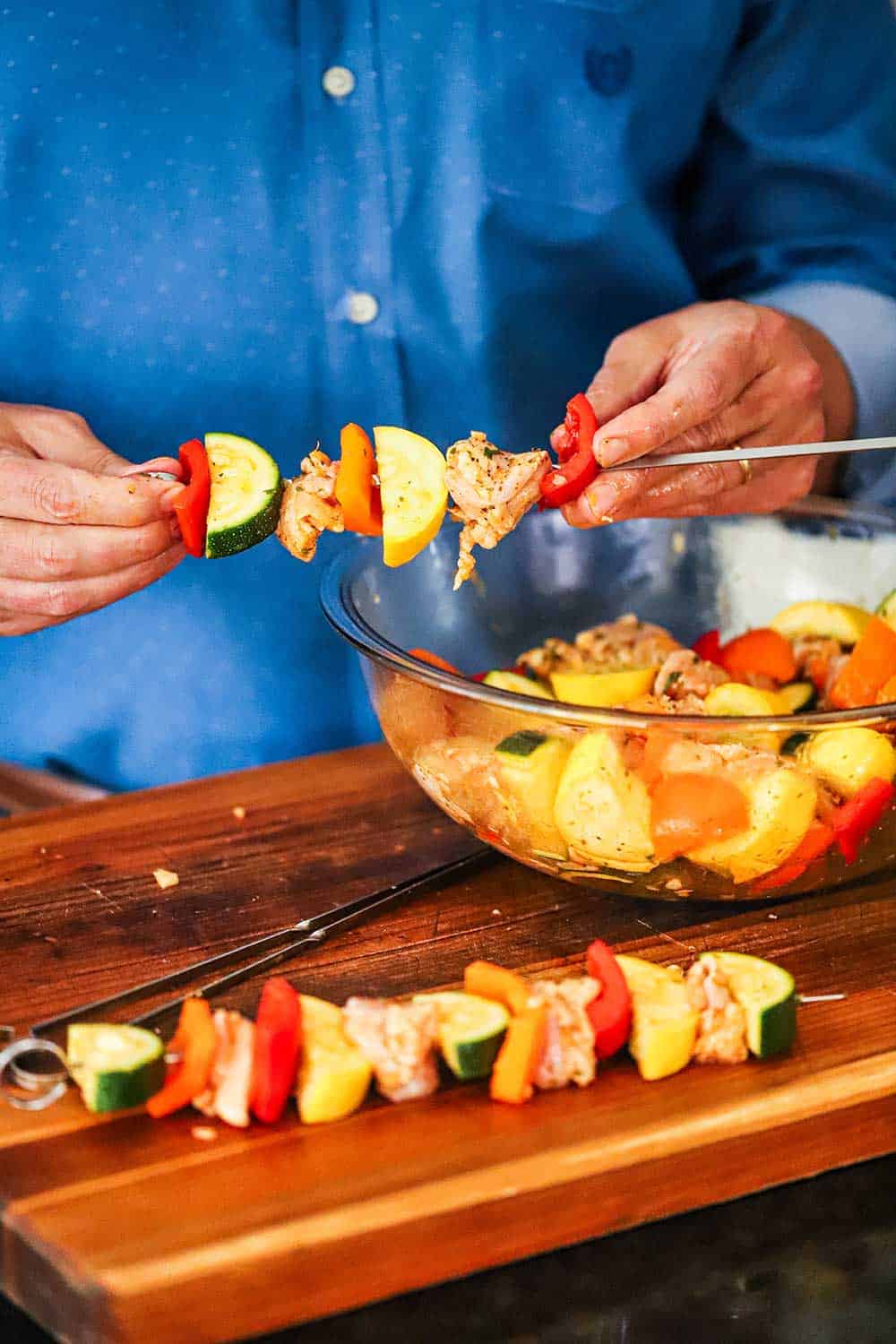 A person in a blue shirt adding pieces of cut chicken and vegetables onto metal skewers.