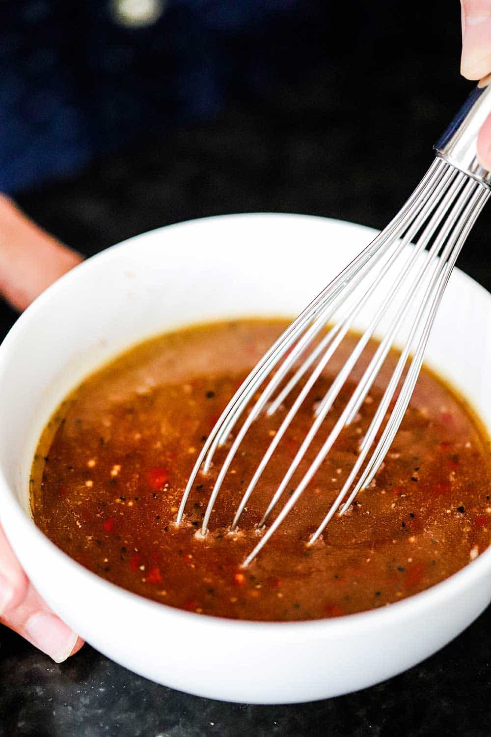 A person holding the handle of a whisk that is inserted into a white bowl filled with a brownish red sauce.