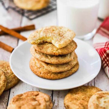 A stack of snickerdoodle cookies on a small white plate surrounded by other cookies and a glass of milk.