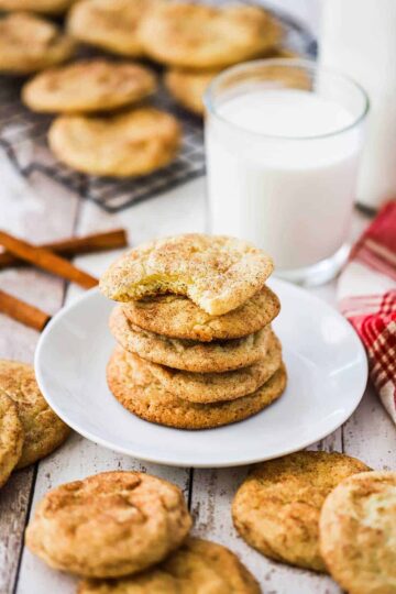 A stack of snickerdoodle cookies on a small white plate surrounded by other cookies and a glass of milk.