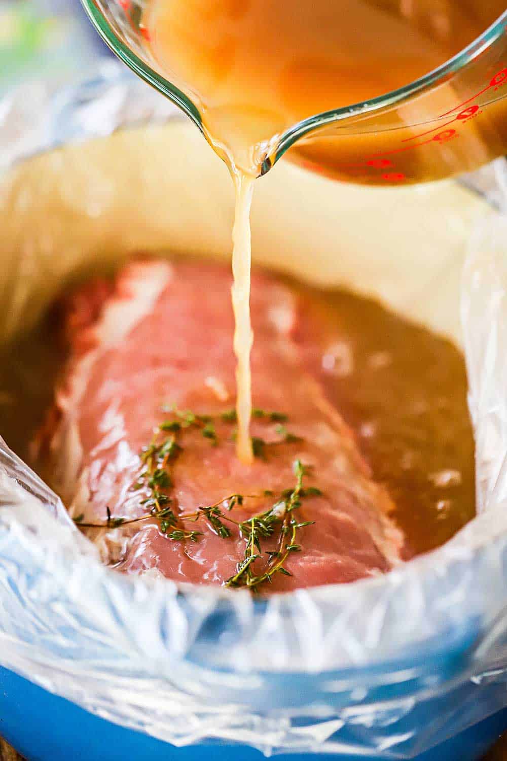 An apple juice brine being poured from a large measuring cup into a Dutch oven lined with a brining bag that has a pork loin in it.
