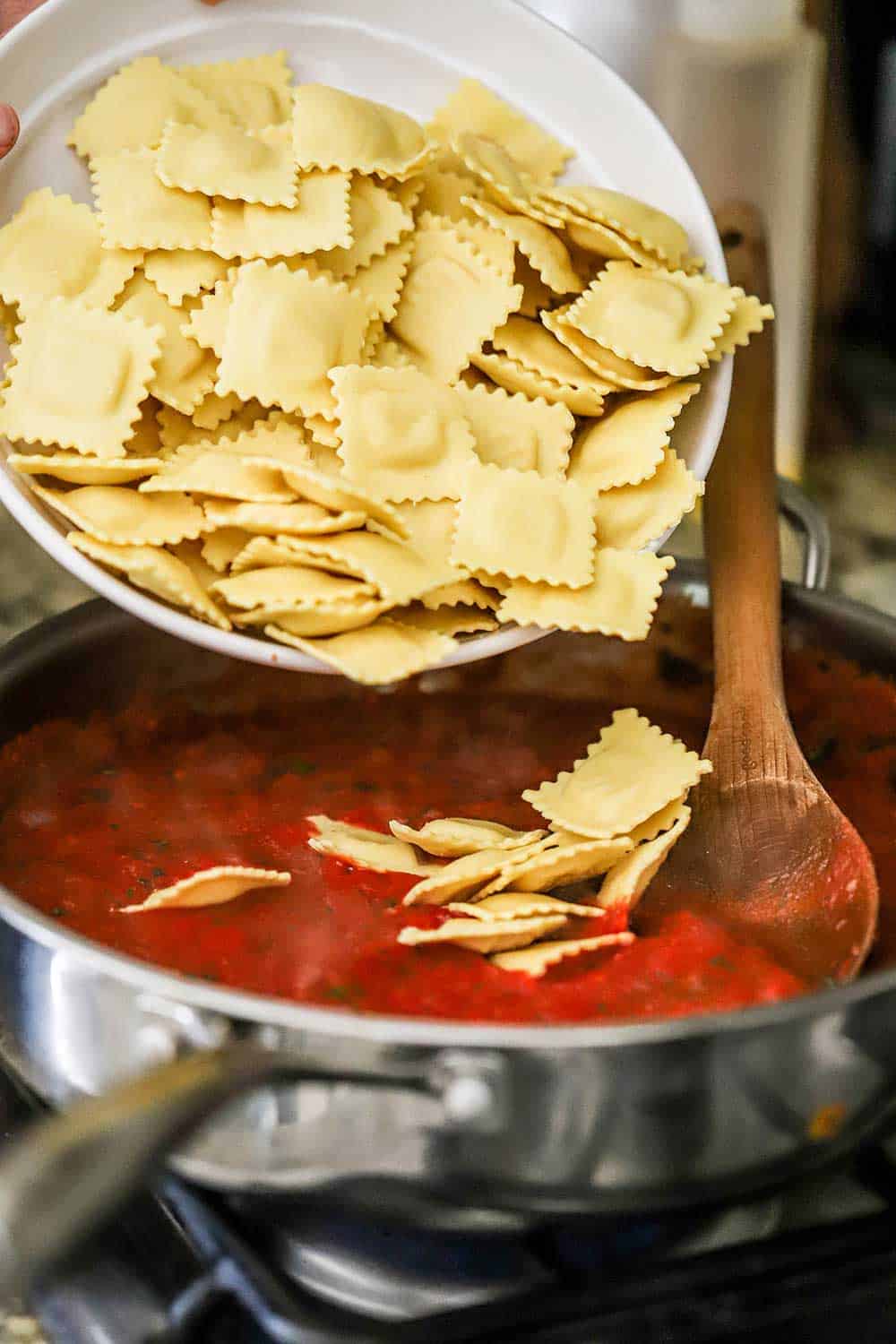 A person dumping a white bowl filled with fresh ravioli into a large skillet filled with a tomato sauce.