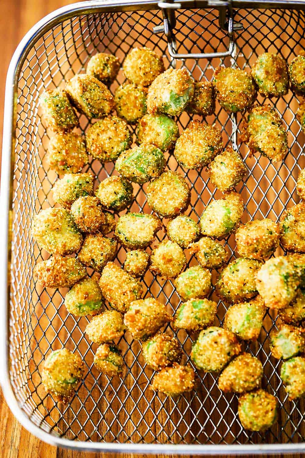 A fryer basket will with fried okra all over a wooden cutting board.