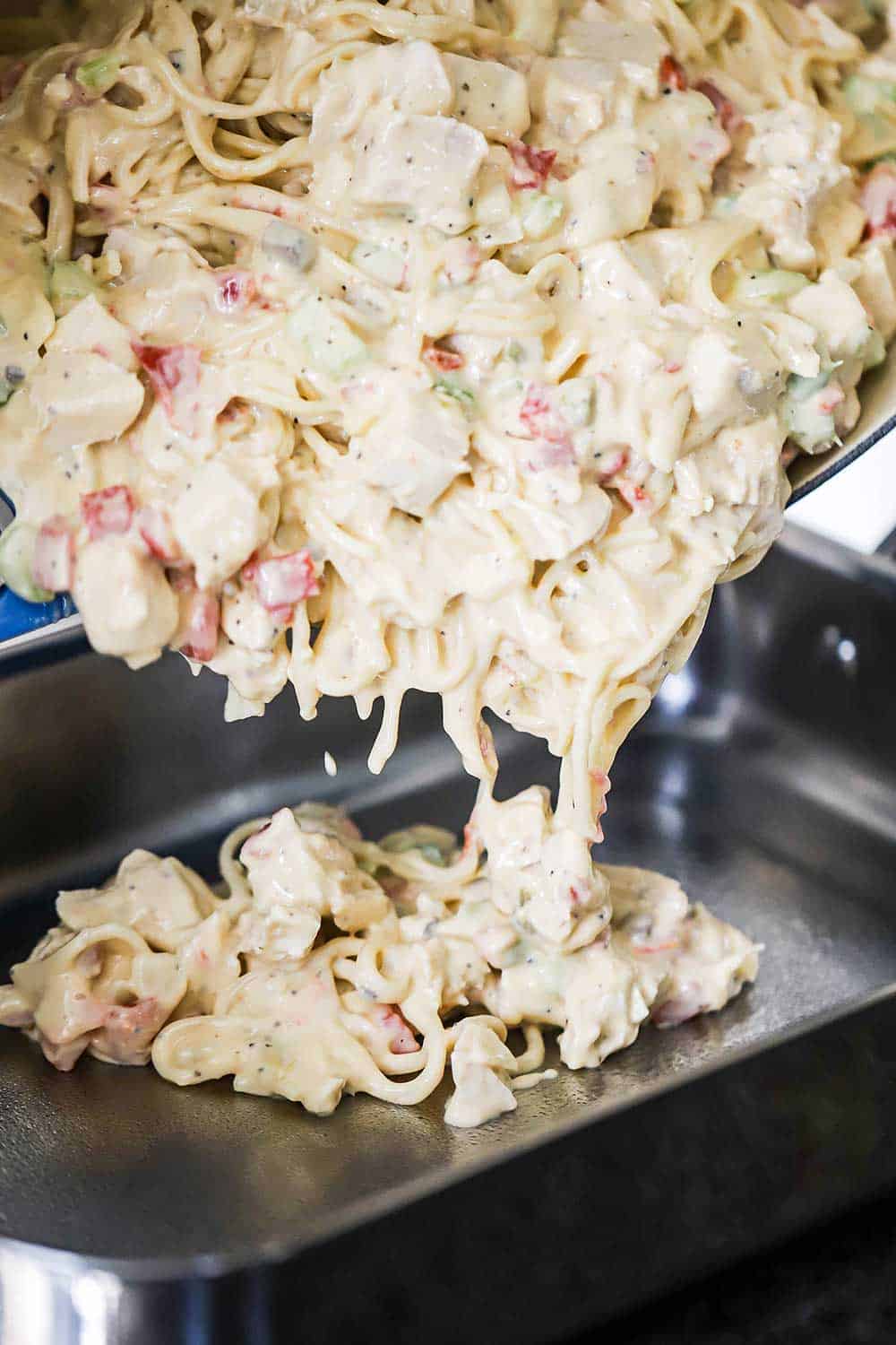 A mixture of cooked spaghetti, chicken, and a melted cheese sauce being poured into a steel lasagna pan.