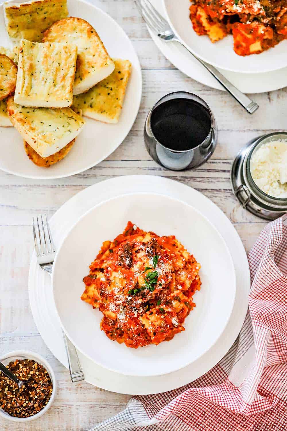 An individual round pasta bowl on a plate filled with easy ravioli and creamy basil-tomato sauce next to a glass of red wine and a plate of sliced garlic bread.