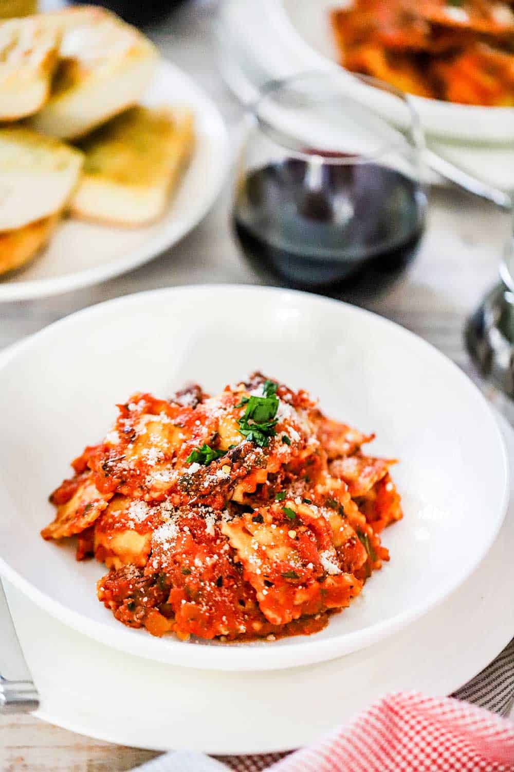An individual white pasta bowl filled with ravioli and a tomato sauce next to a stemless wine glass filled with red wine and a plate of garlic bread nearby.