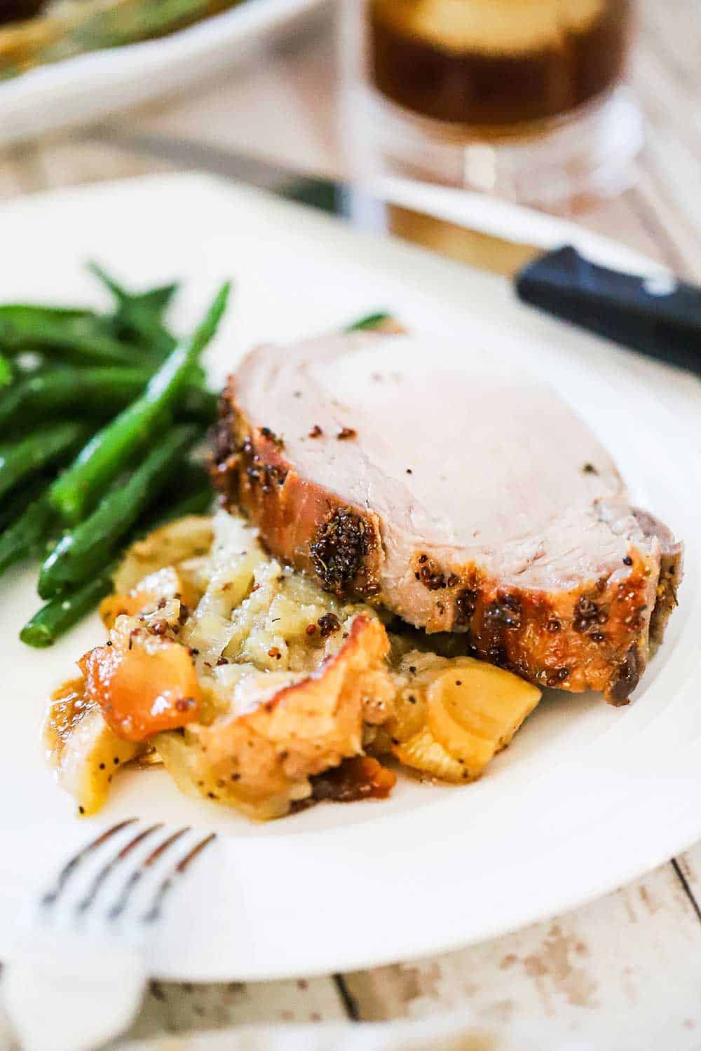 A white dinner plate filled with a slice of roasted pork loin and roasted apples and fennel and steamed green beans.