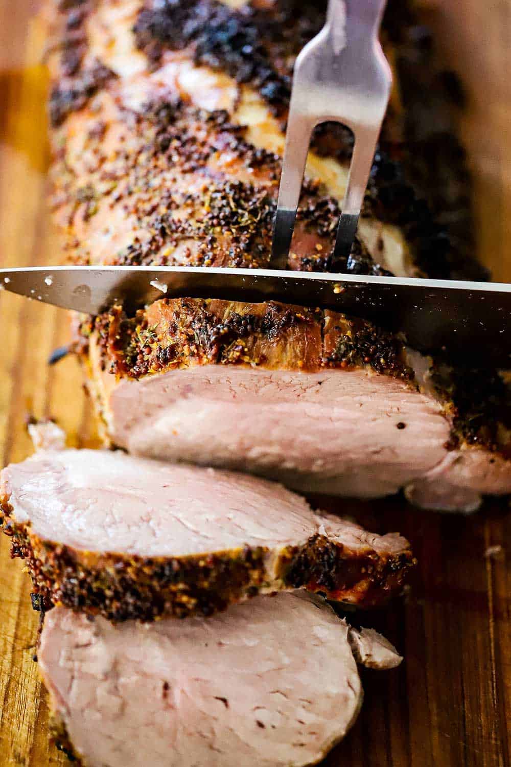 A large butcher knife slicing through a large roasted pork loin roast on a wooden cutting board.