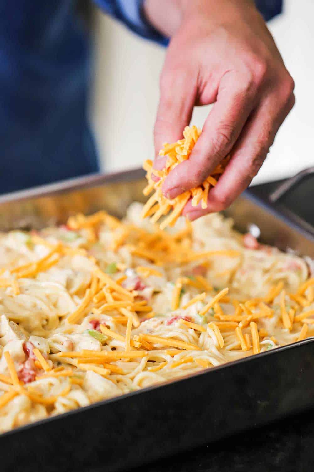 A hand sprinkling shredded cheddar over the top of an uncooked pan of chicken spaghetti.