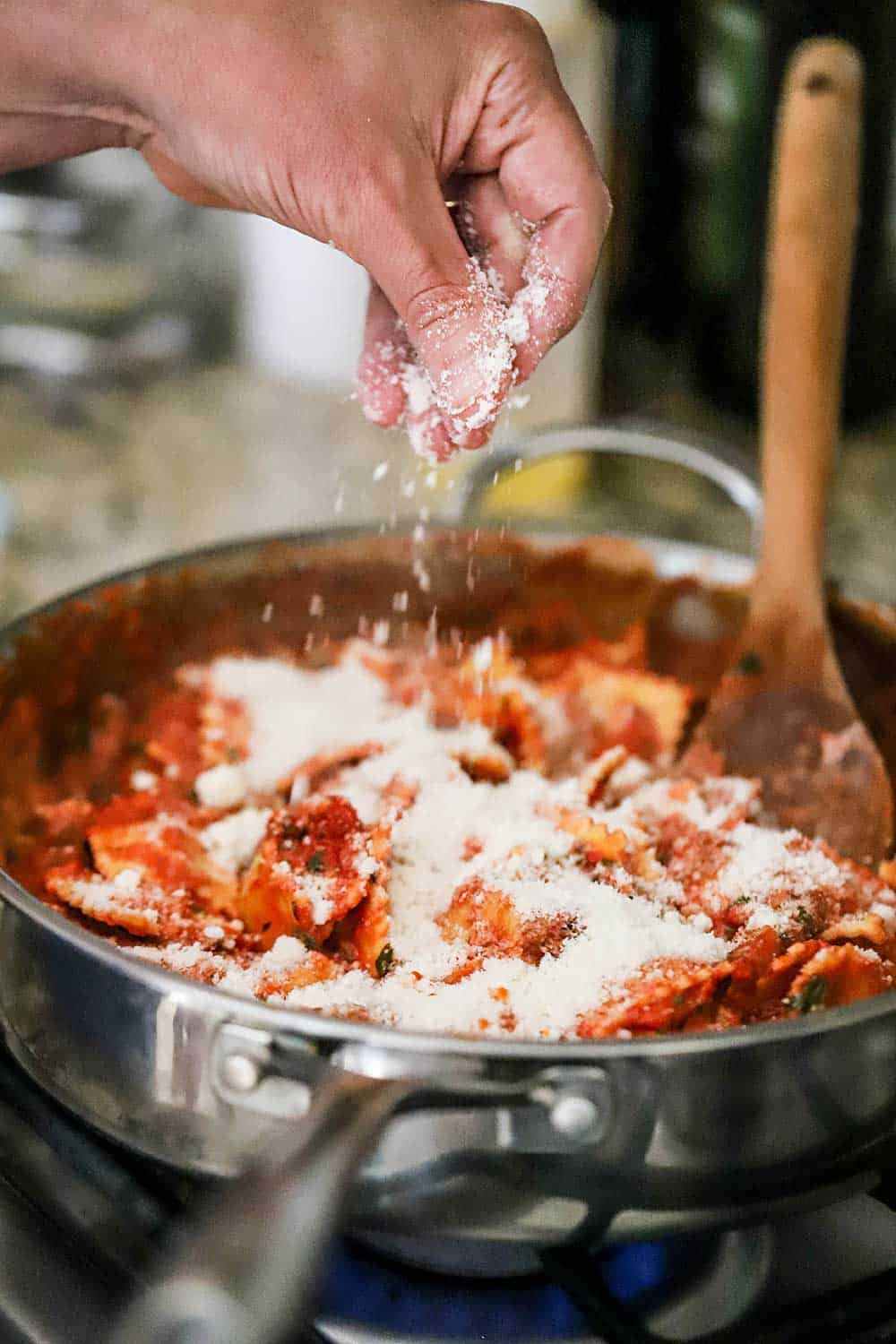 A person sprinkling grated parmesan cheese into a large silver skillet filled with ravioli in a tomato sauce.