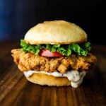 A chicken fried steak sandwich topped with sliced tomato and lettuce sitting on a wooden cutting board.