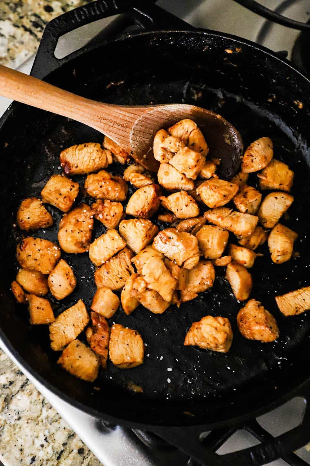 Chicken pieces in a large cast-iron skillet being stirred by a wooden spoon.