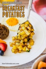 A silver serving spoon transferring a helping of breakfast potatoes onto a white plate with an egg, fruit, and sausage patty on it.