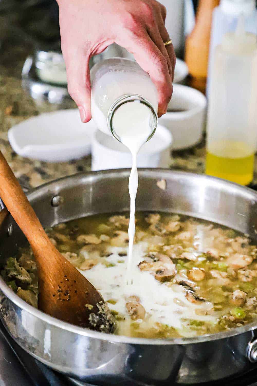 A person pouring milk from a small milk jug into a skillet filled with sautéed mushrooms and chicken stock.