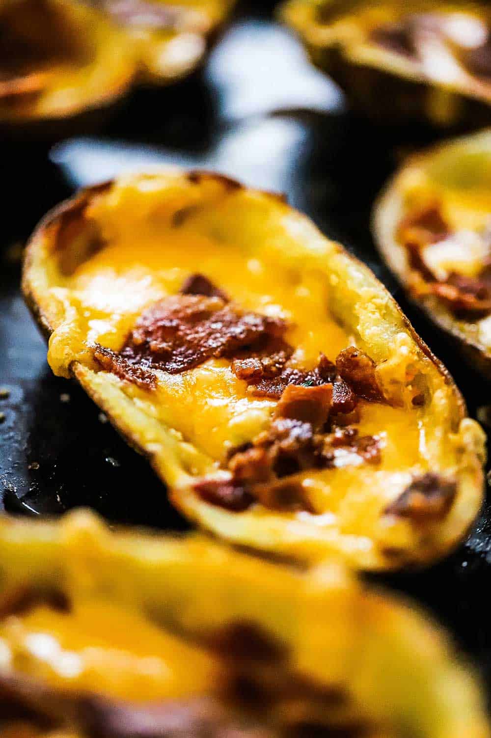 A potato skin on a baking sheet filled with melted cheddar cheese and topped with cooked crumble bacon.