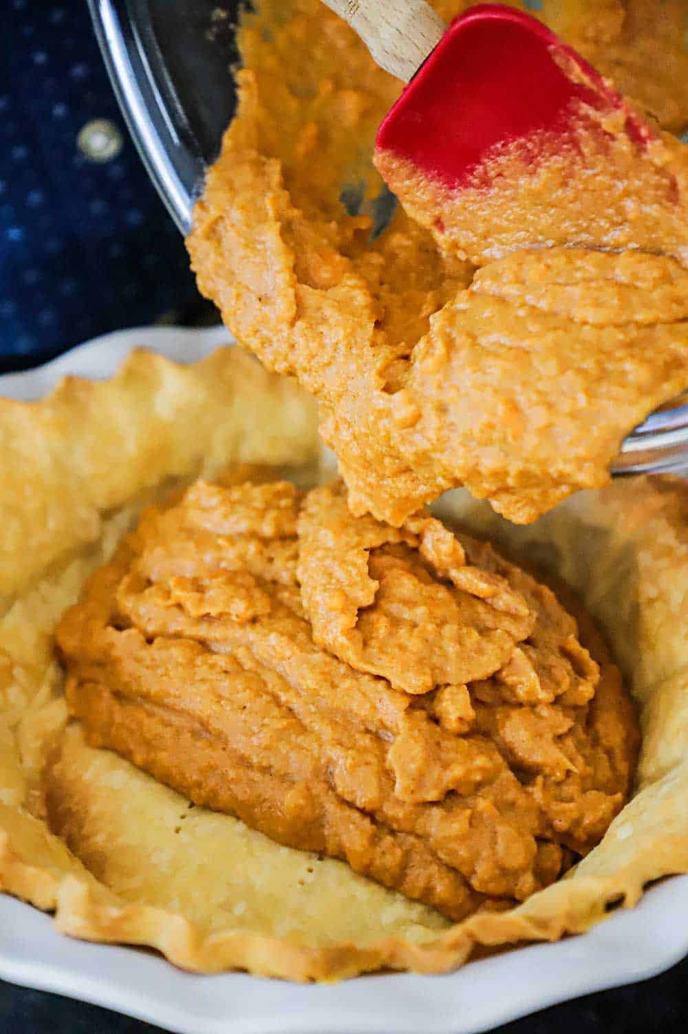 A person transferring sweet potato pie filling from a large glass bowl into a pie crust-lined pie dish.