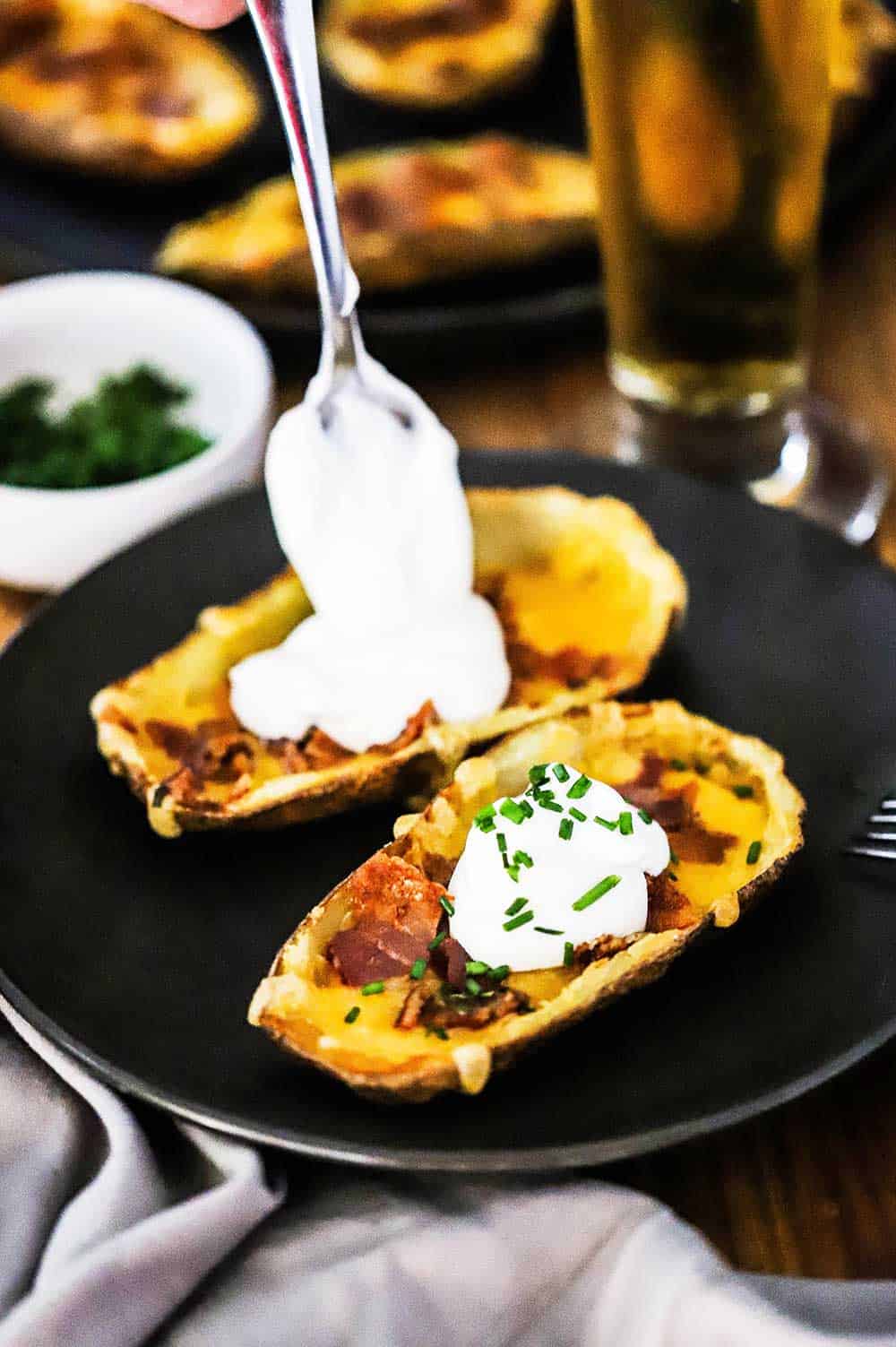 A person adding sour cream on top of loaded potato skins sitting on a black plate.