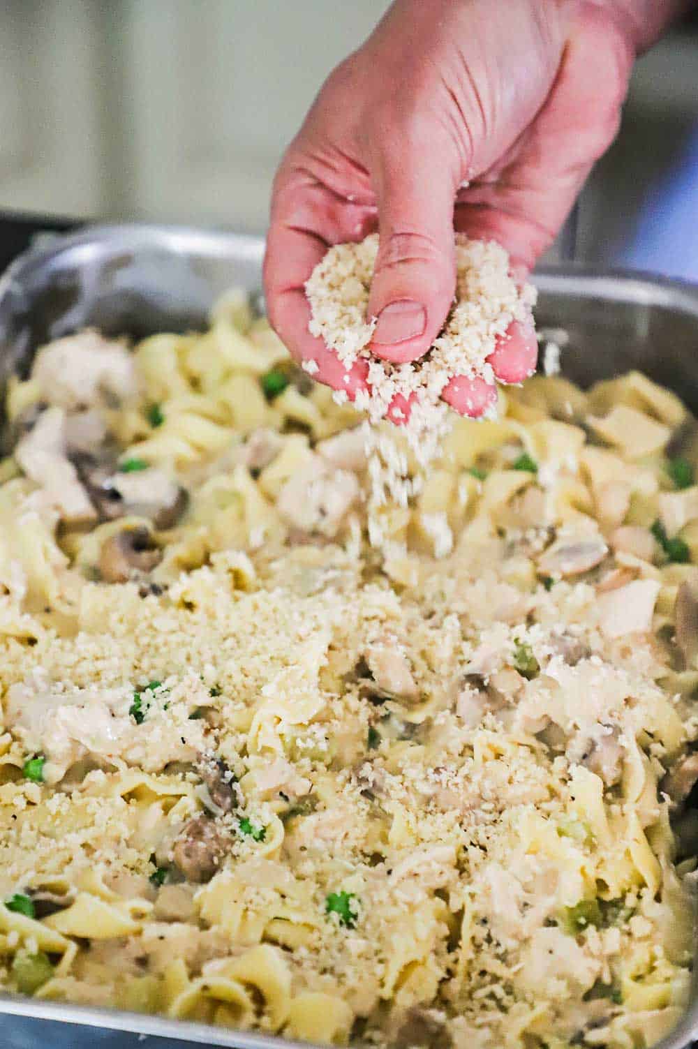A hand sprinkling breadcrumbs over the top of turkey tetrazzini in a lasagna pan.