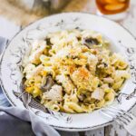 An antique serving bowl filled with a large helping of turkey tetrazzini sitting next to a glass of iced tea.