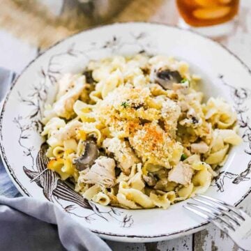 An antique serving bowl filled with a large helping of turkey tetrazzini sitting next to a glass of iced tea.