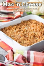 Mac n cheese with roasted cauliflower and smoked bacon in a white dish next to a red patterned napkin with a spoon on top of a patterned place mat