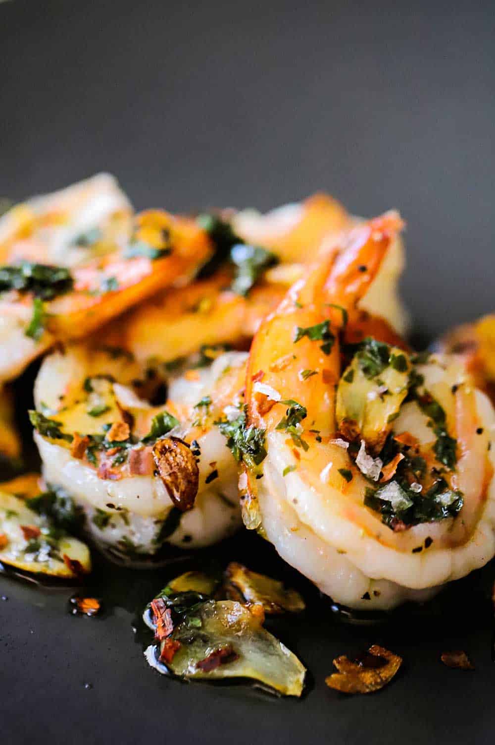 A small dark grey appetizer plate filled with 4 cooked shrimps topped with fried garlic slivers and a white wine and parsley sauce.