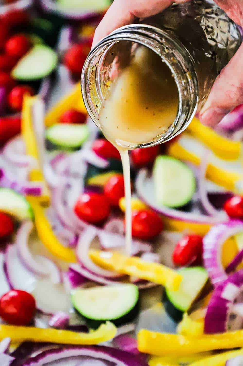 A person pouring a Greek dressing from a small jar onto cut vegetables spread out on a sheet pan.