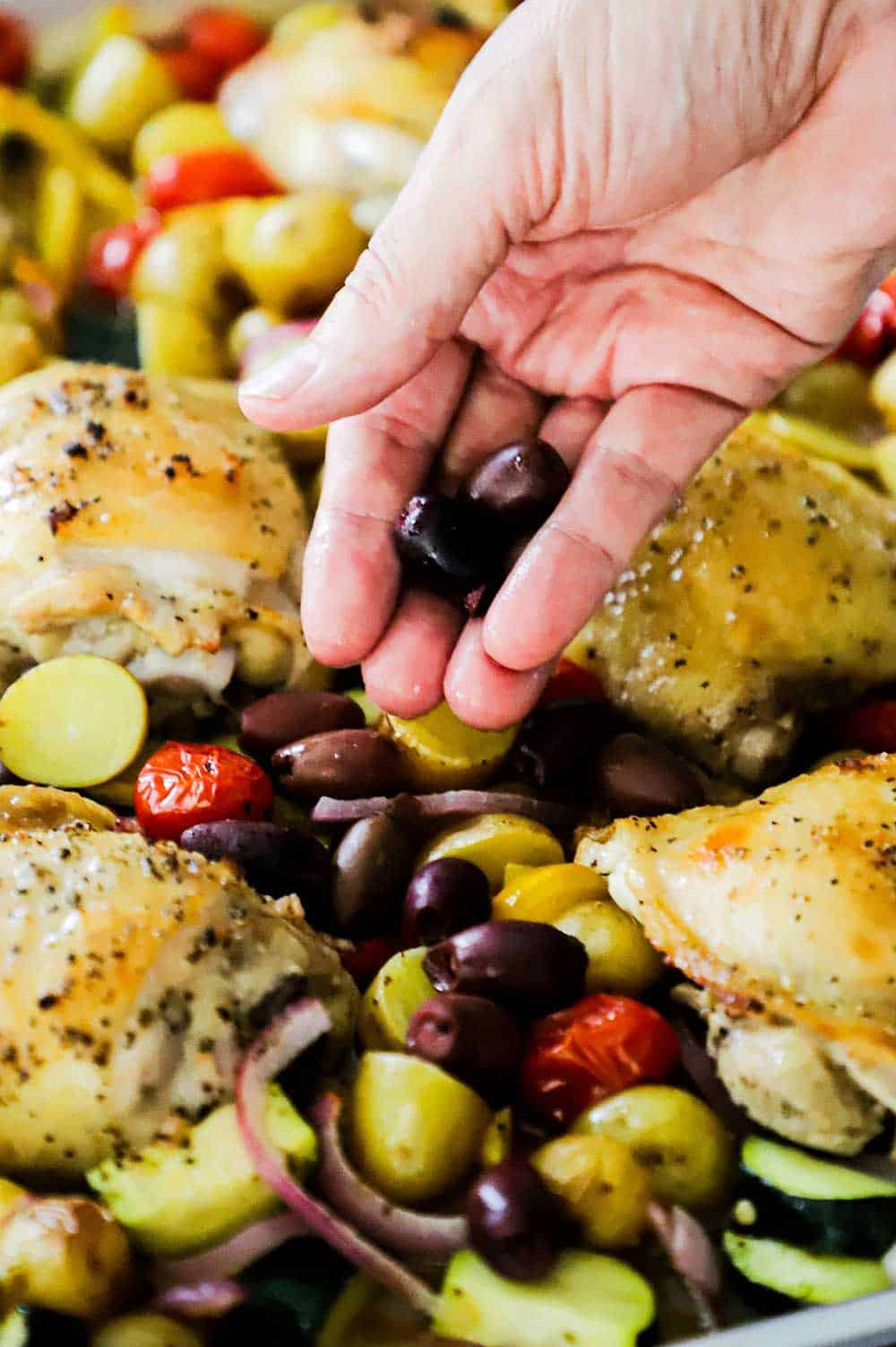 A person's hand sprinkling Kalamatas olives on the top of sheet pan Greek chicken.
