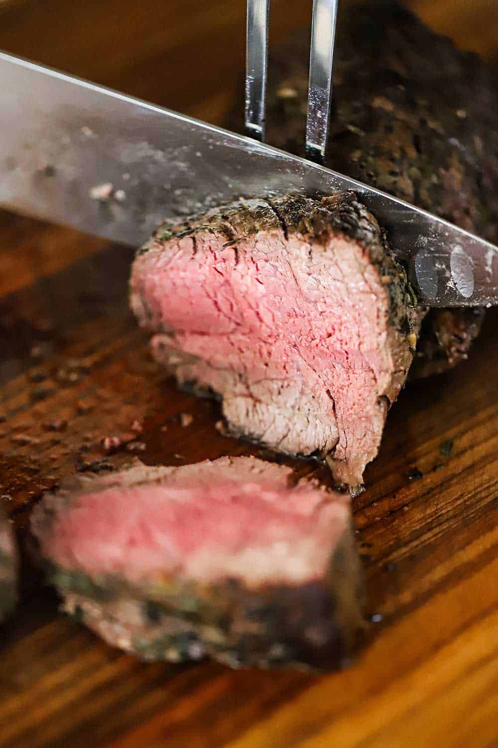 A large meat fork piercing a beef tenderloin roast with a knife cutting away slices from it.