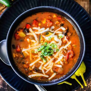 A blue soup bowl filled with vegetable chili and black beans next to a red bell pepper and a bunch of cilantro.