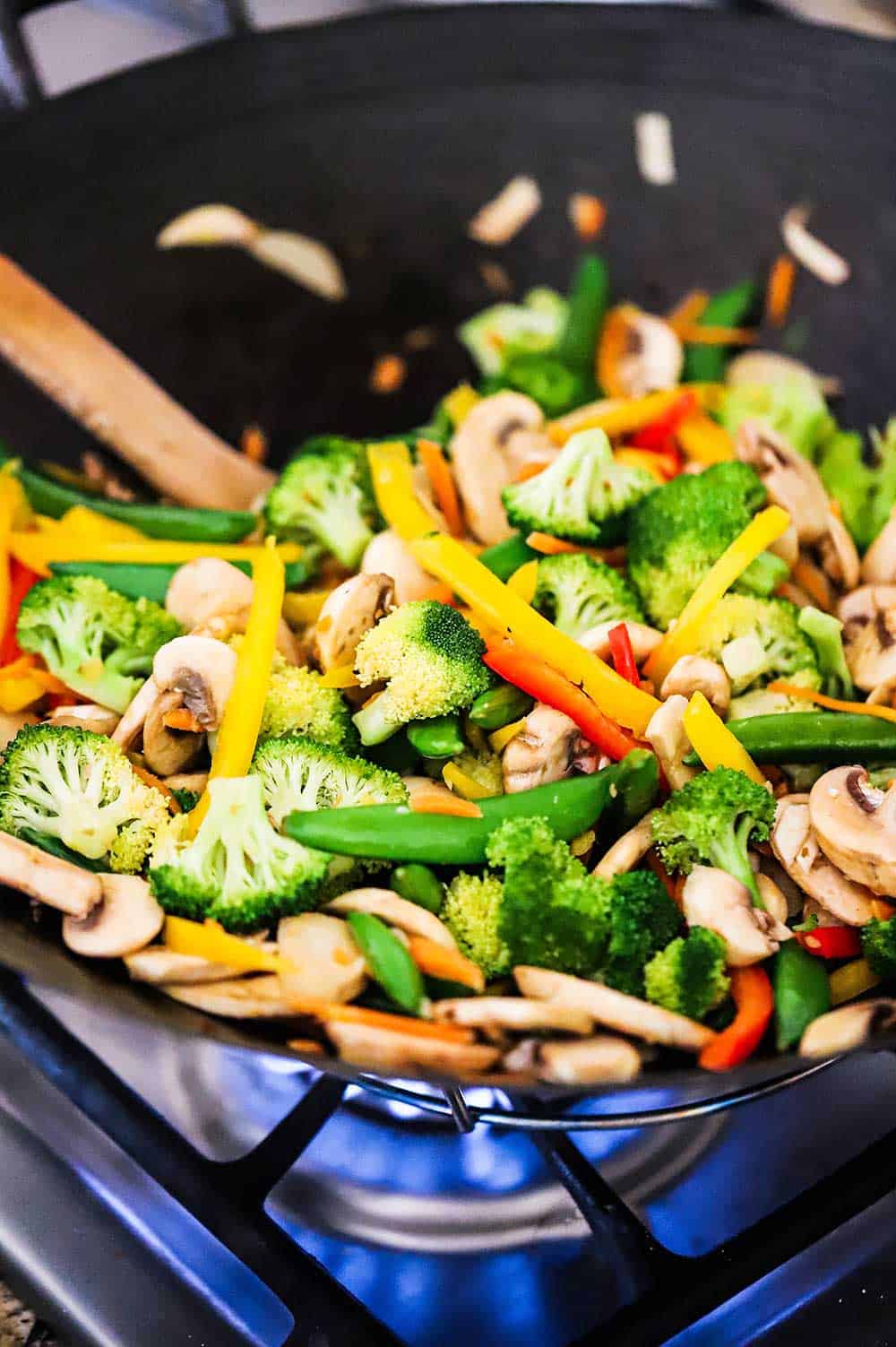 A large black wok over a gas flame on a stove with vegetable stir-fry cooking inside it.