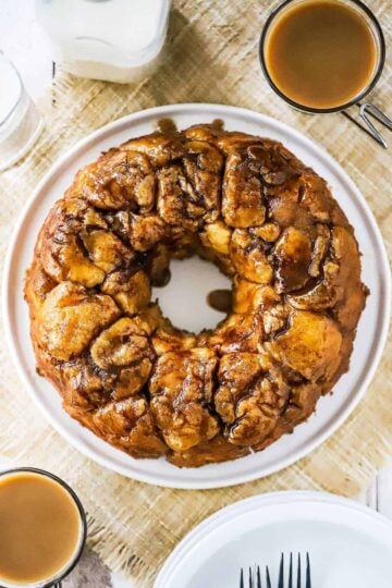 An untouched circular loaf of homemade monkey bread on a circular white plate next to a couple mugs of coffee.