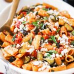 A large white oval serving platter filled with a tomato pasta topped with cooked eggplant pieces and crumbled cheese.
