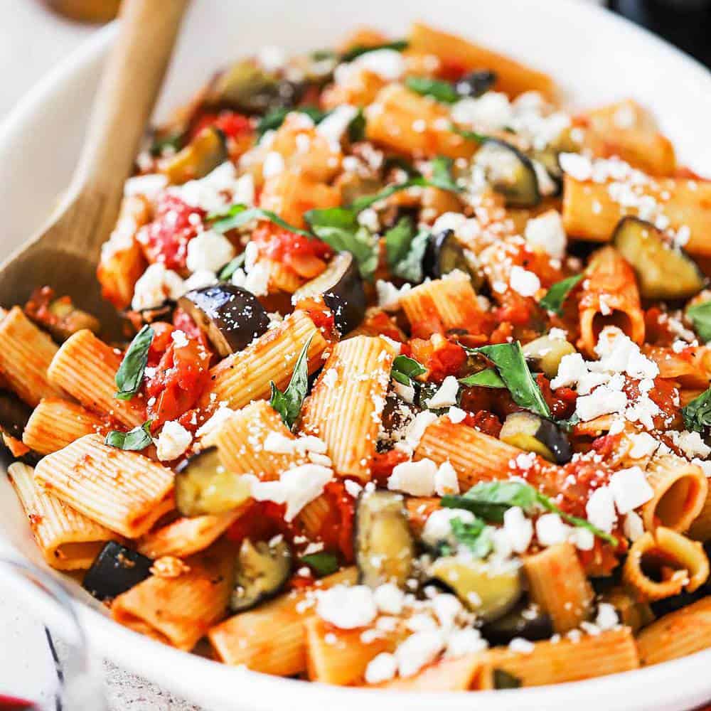A large white oval serving platter filled with a tomato pasta topped with cooked eggplant pieces and crumbled cheese.