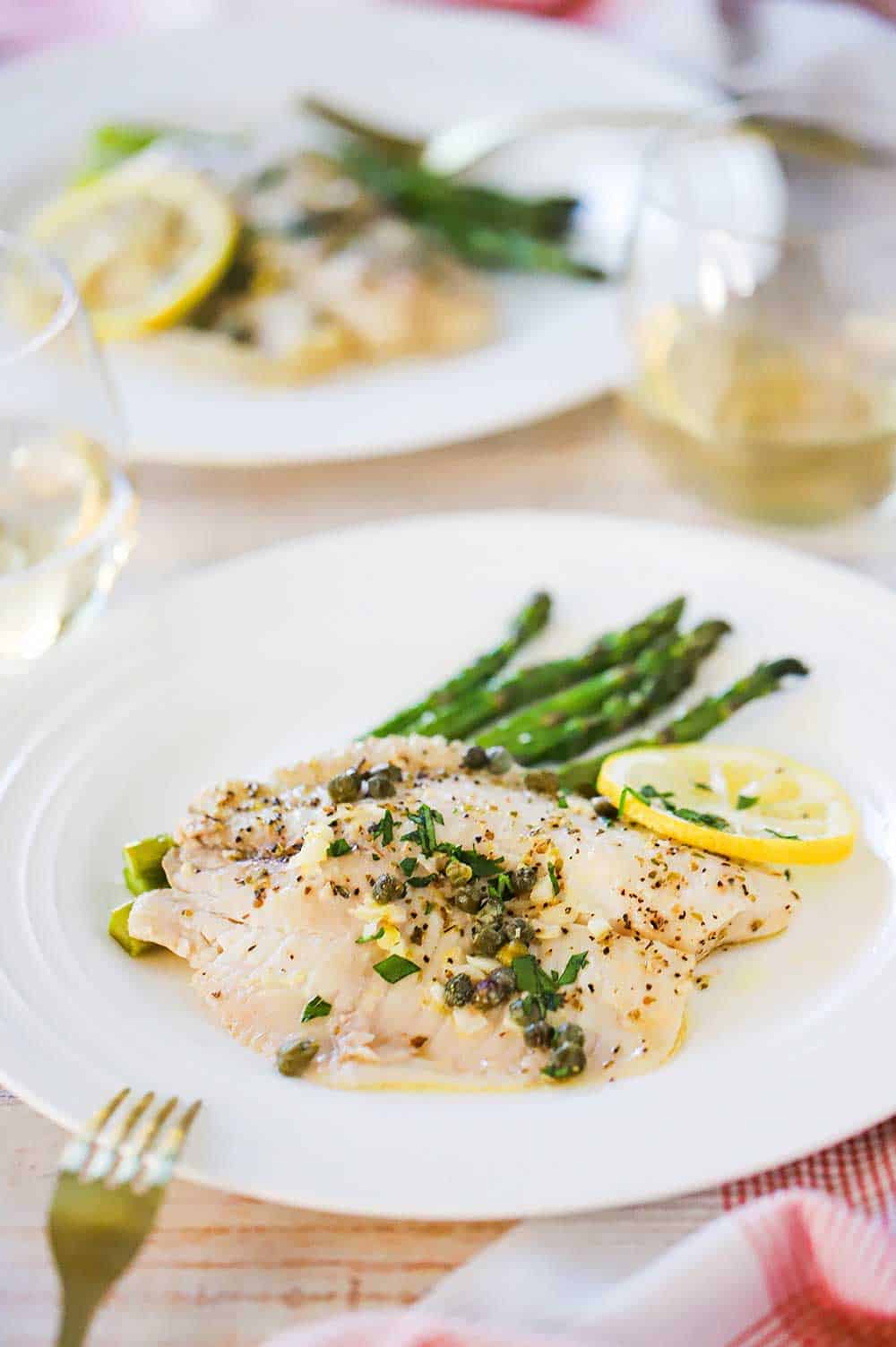 Two individual dinner plates filled with baked tilapia with lemon garlic sauce sitting next to roasted asparagus and a lemon wedge.