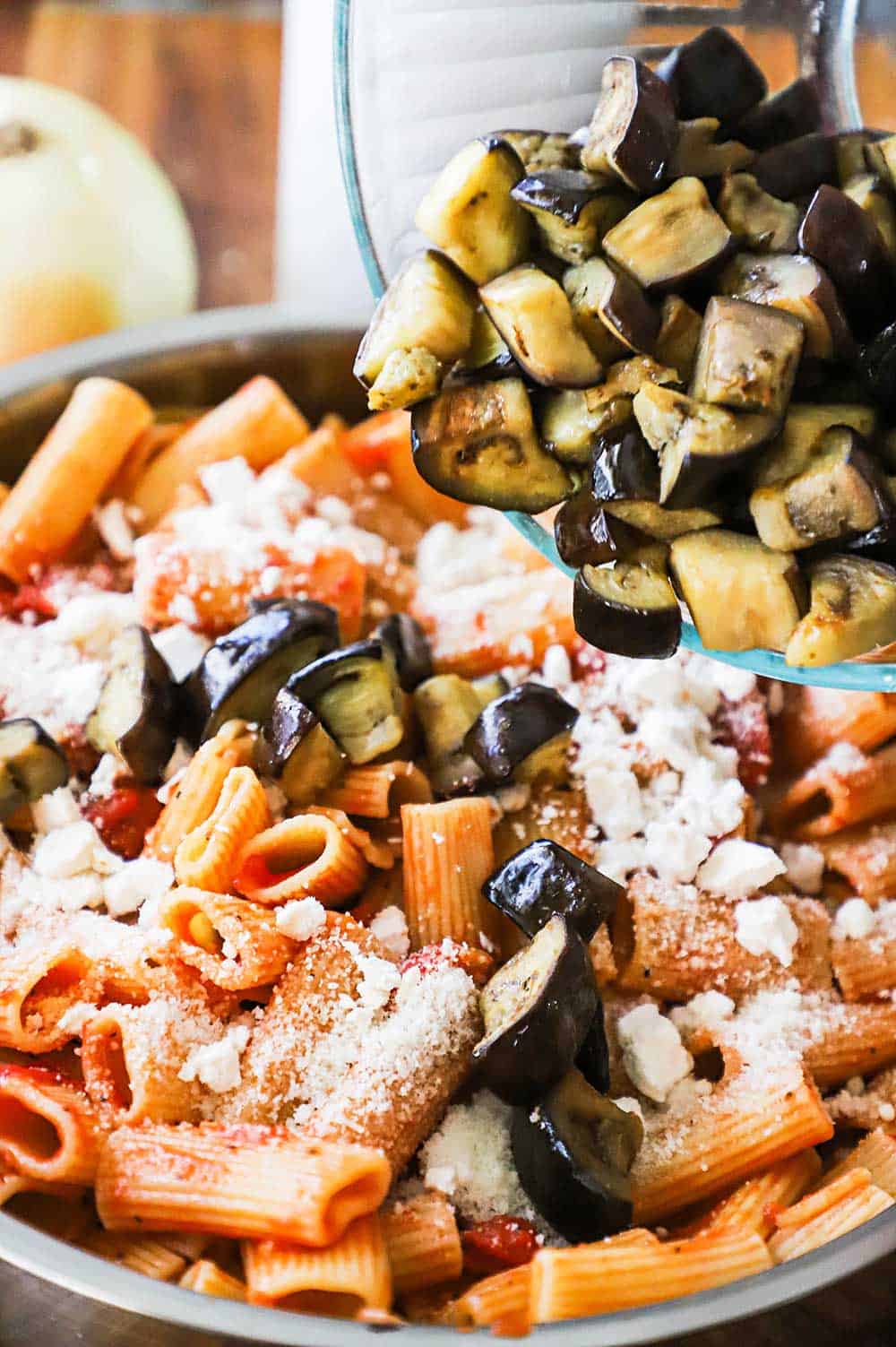 Pieces of sautéed eggplant being transferred from a glass bowl into a large skillet filled with cooked rigatoni pasta and shredded ricotta selata cheese.