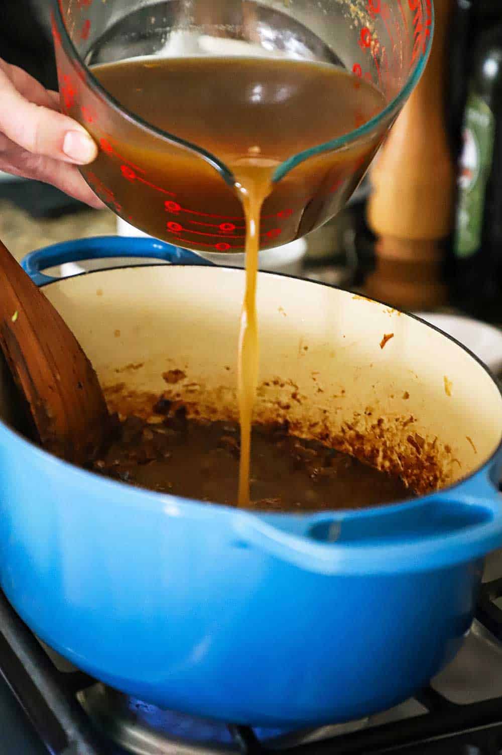 A person pouring beef stock into a blue Dutch oven filled with caramelized onions.