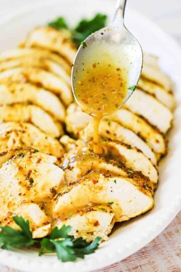 A spoon pouring a honey mustard glaze over slices of baked chicken arranged on a white oval serving platter.
