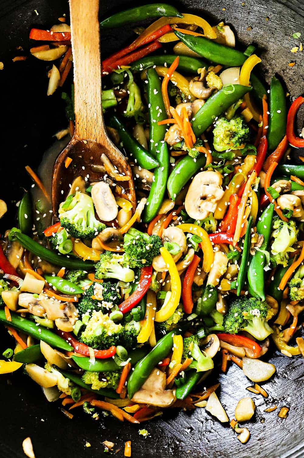 A black metal wok filled with vegetable stir-fry with a wooden spoon stuck in the side.