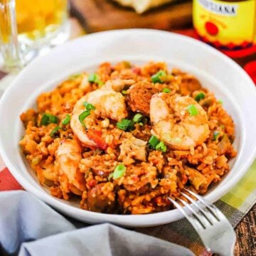 A white dinner bowl filled with shrimp and sausage jambalaya next to a bottle of Louisiana hot sauce and a mug of beer.