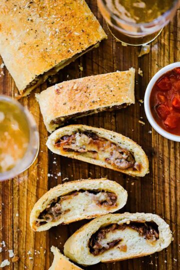 Four slices of sausage bread sitting next to the cut loaf of the bread all next two a bowl of marinara and two glasses of beer.