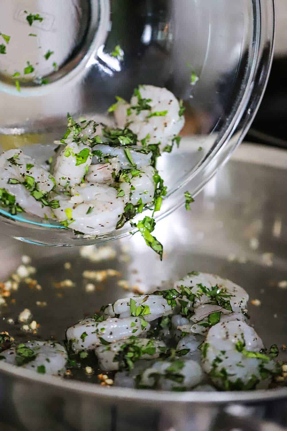 Uncooked shrimp that has been coated with fresh herbs being transferred into a skillet filled with sautéed garlic.