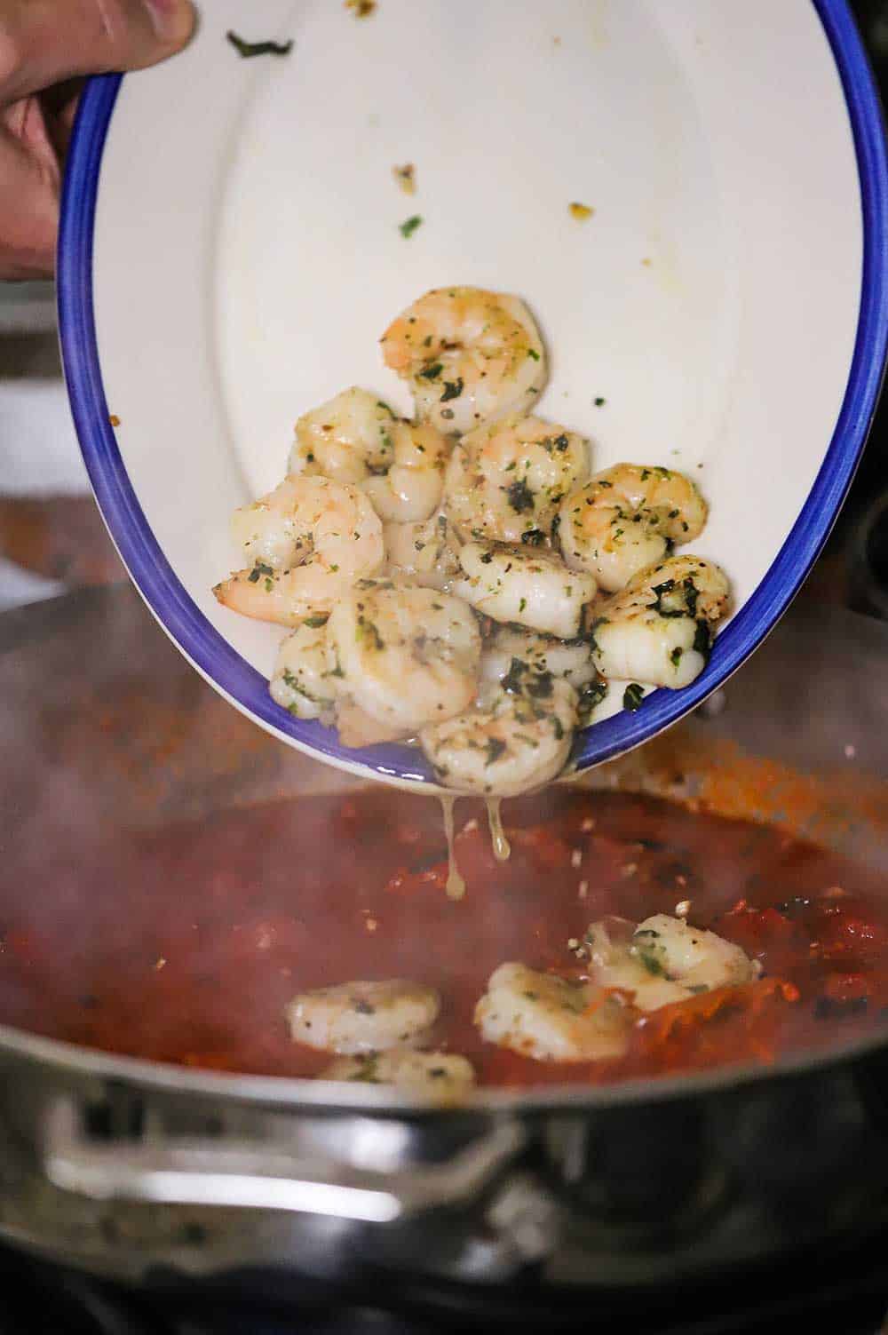 Sautéed shrimp being transferred from a platter into a skillet filled with simmering marinara sauce.