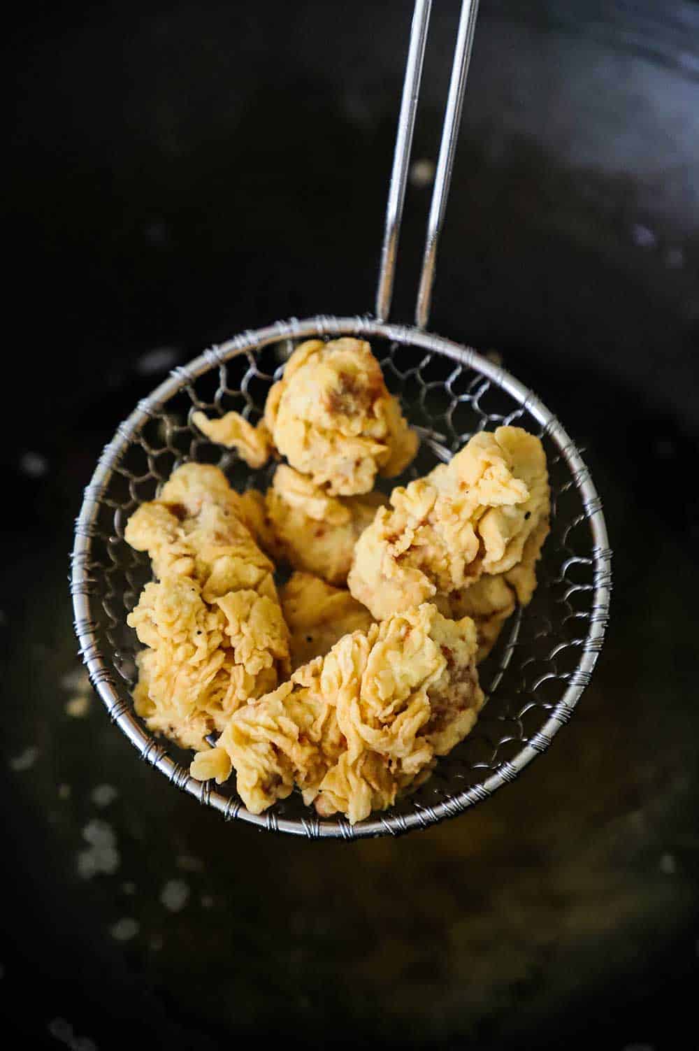 A kitchen spider holding up pieces of fried chicken nuggets over a wok filled with hot oil.