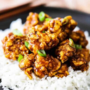 A black dinner plate filled with white rice topped with orange chicken and garnished with chopped scallions.