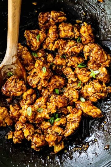 An overhead view of a wok filled with orange chicken with a wooden spoon inserted in the side.