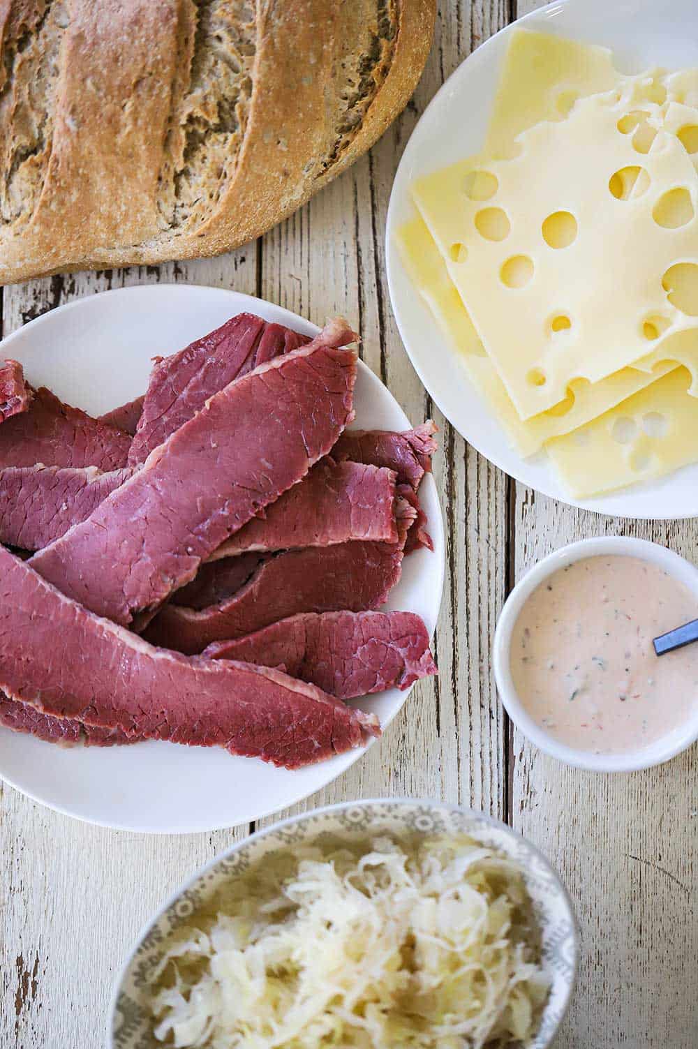 An overhead view of a loaf of rye bread, a plate of sliced Swiss cheese, a plate of sliced cooked corned beef, a small bowl of Russian dressing, and a bowl of sauerkraut.