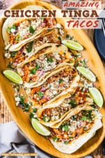 A large oval wooden platter that is filled with six chicken tinga tacos with cut limes surrounding them and crumbled cheese sprinkled over the top.