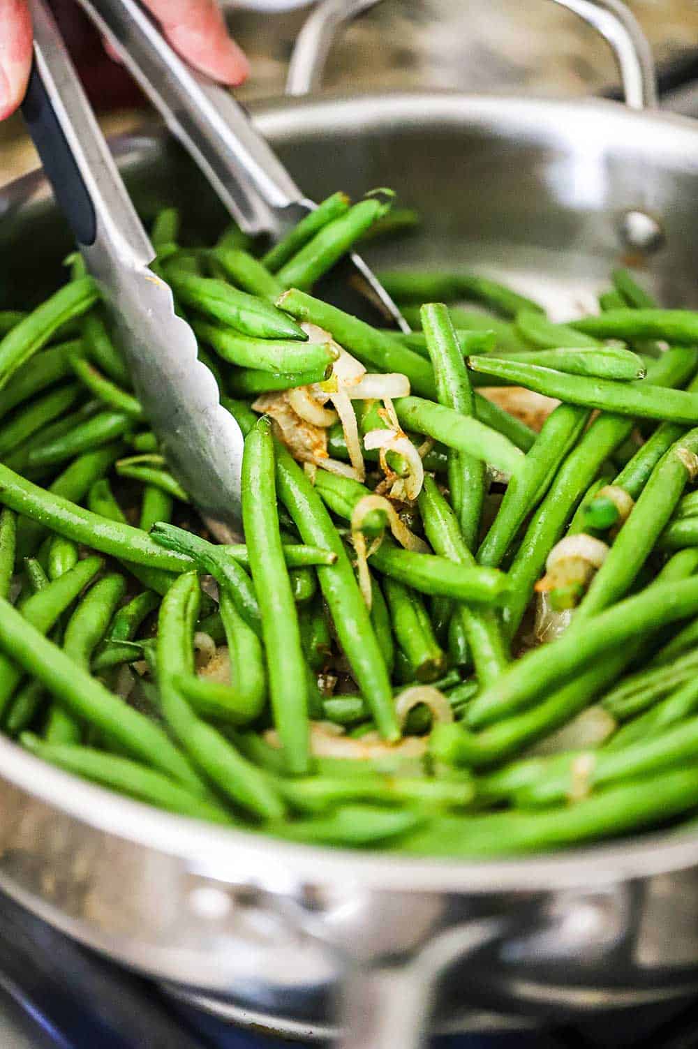A person using a metal spatula to stir fresh green beans with sautéd onions in a large silver skillet.