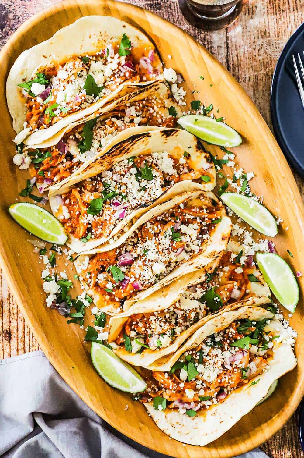 A large oval wooden platter that is filled with six chicken tinga tacos with cut limes surrounding them and crumbled cheese sprinkled over the top.